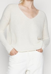 Bílý, měkký, plyšový svetr s výstřihem do V a dlouhými rukávy, s volným střihem a texturovanou tkaninou. V kombinaci s bílými kalhotami.