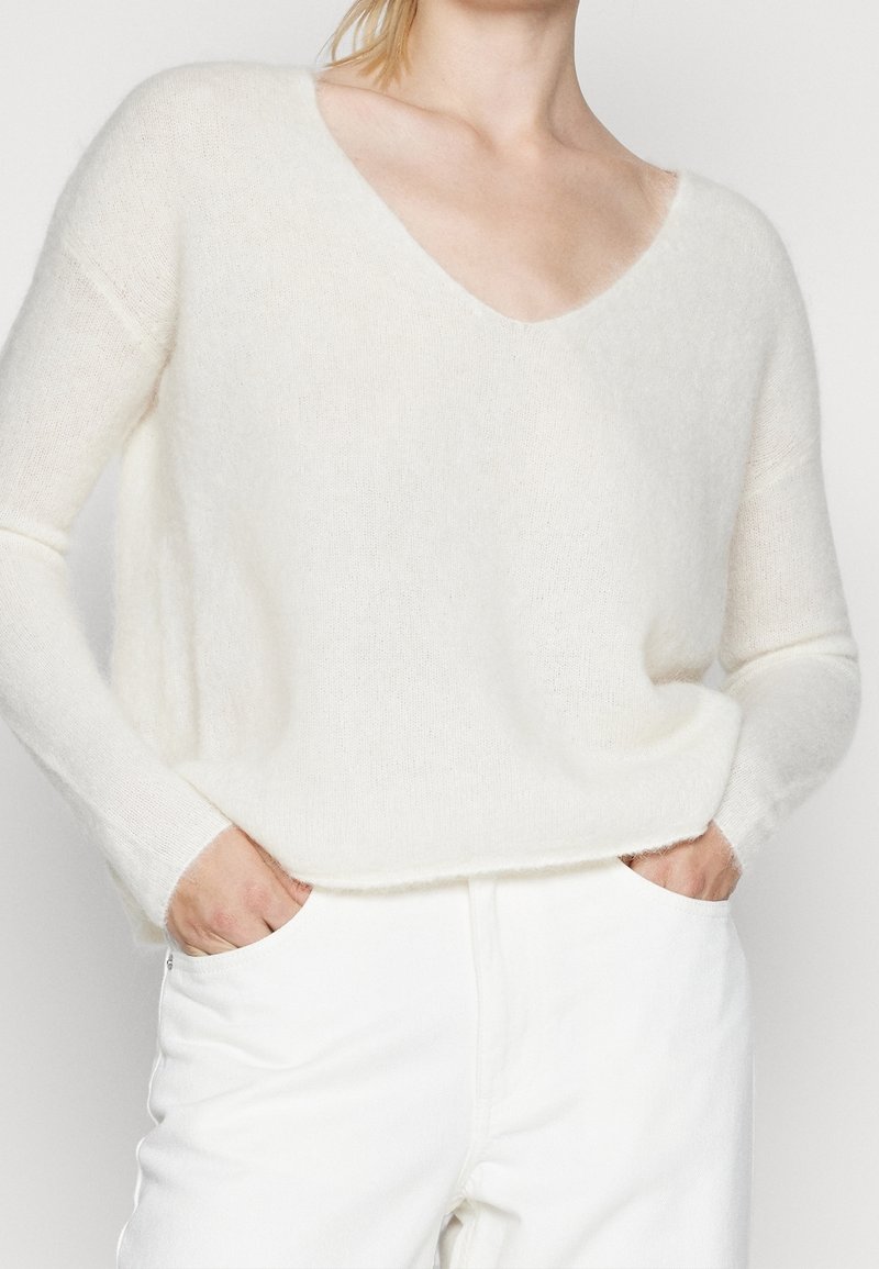 Bílý, měkký, plyšový svetr s výstřihem do V a dlouhými rukávy, s volným střihem a texturovanou tkaninou. V kombinaci s bílými kalhotami.