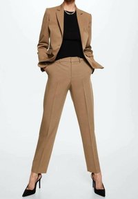 Personne portant un costume beige sur mesure avec un haut noir, les mains dans les poches, et des escarpins à bouts pointus noirs, se tenant jambes écartées.