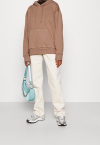 Sweat à capuche marron avec une poche avant, associé à un pantalon écru et des baskets argentées. Tenant un sac à main bleu clair avec un détail en chaîne.