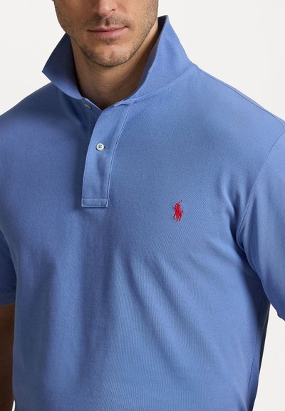 Plava polo majica izrađena od teksturiranog materijala. Ima okrenutu kragnu, dva dugmeta i crveni izvezeni logo na lijevoj strani prsnog koša.