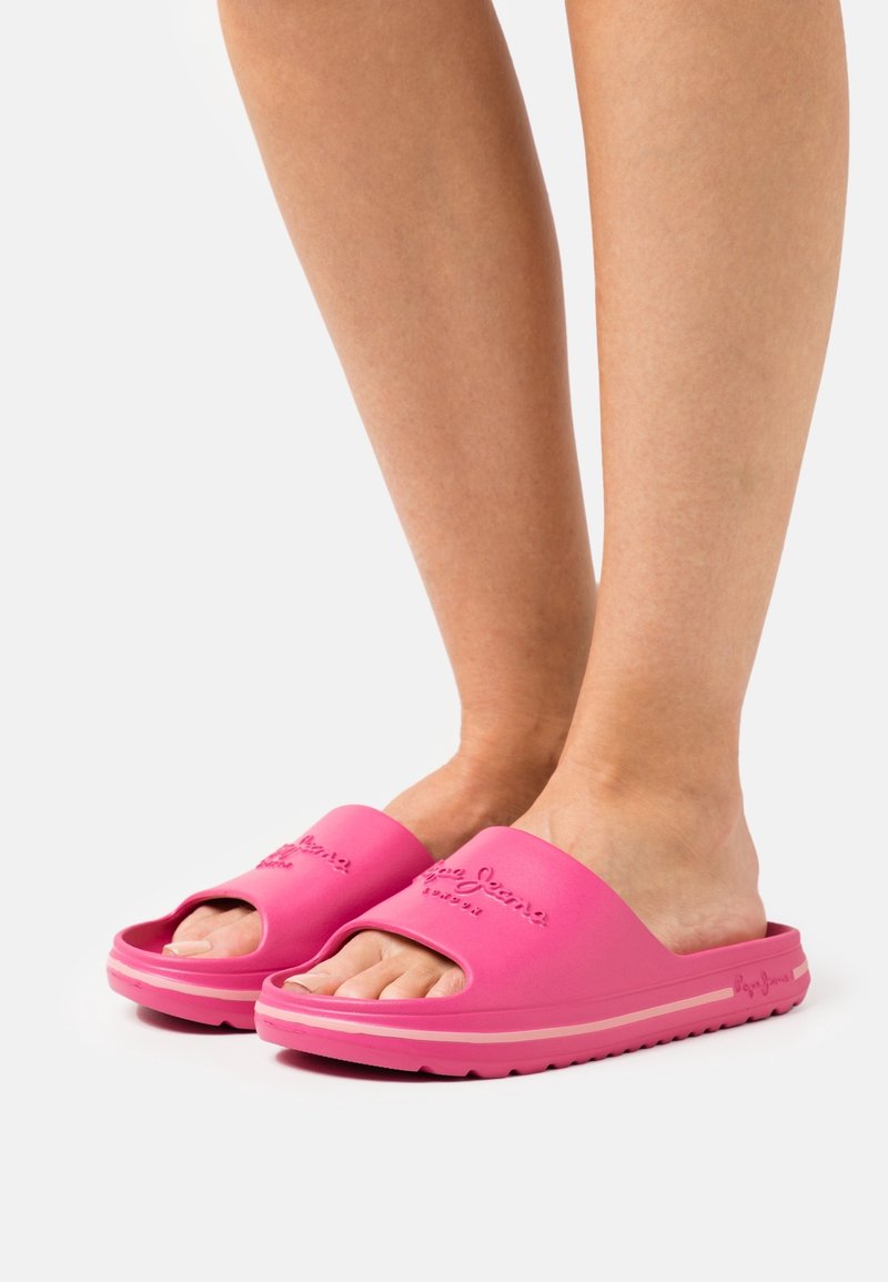 Pieds portant des sandales roses vives à enfiler avec une semelle texturée et un logo en relief sur la lanière, sur fond blanc.
