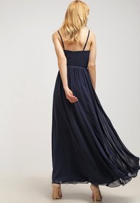 Žena s blond vlasy, oblečená v volném námořnickém modrém maxi šatech bez rukávů a béžových vysokých podpatcích, stojí zády k pozorovateli.