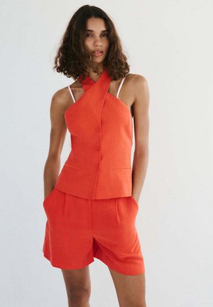 Mujer con cabello rizado que lleva un top naranja sin mangas con botones y shorts de talle alto a juego con las manos en los bolsillos.
