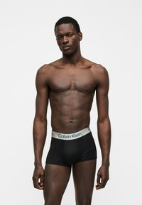 Homme musclé debout devant un fond uni portant un boxer Calvin Klein noir avec ceinture blanche.