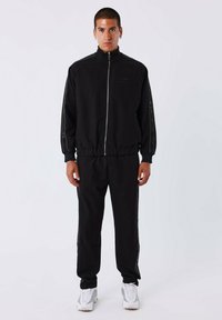 Tuta nera con giacca con zip e vita elasticizzata. Presenta bande laterali con dettagli in rete. Indossato con scarpe sportive bianche.