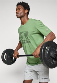 Grünes Baumwoll-T-Shirt mit einem schwarzen Champion-Logo, kombiniert mit grauen Sportshorts, während er eine Langhantel mit einer schwarzen Gewichtscheibe hält.