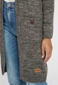 Gestrickter langer Cardigan in Grau mit einem strukturierten Muster, ausgestattet mit einem einzelnen Knopf, seitlichen Taschen und einem braunen Logotag am Saum.