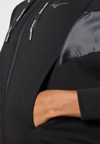 Felpa nera con zip, dal tessuto morbido, con accenti neri lucidi sulle spalle e una mano nella tasca laterale. Dettagli in metallo argentato.