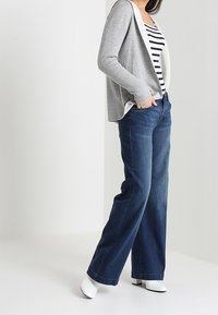 Kvinna klädd i en grå cardigan, svart-vit randig skjorta, vida blå jeans och vita ankelboots, stående med händerna i fickorna.