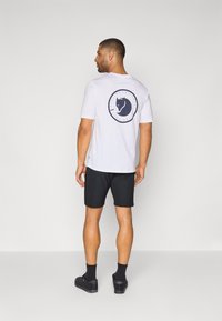 Weißes T-Shirt mit einer runden navyblauen Grafik, die den Kopf eines Pferdes auf dem Rücken zeigt. Getragen mit schwarzen Shorts und schwarzen Schuhen.