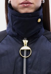 Μπλε σ ακάκι με ψηλό γιακά, φερμουάρ με χρυσά στοιχεία και κυκλικό τζαμάκι, καθώς και έντονες ραφές.