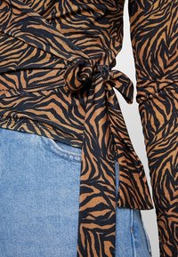 Braun- und schwarz gestreiftes Wickeltop mit einer Bindeschließe, aus einem glatten Stoff gefertigt. Kombiniert mit hellblauer Jeans.
