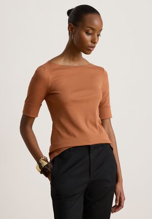 Femme à la peau foncée portant un top à manches courtes couleur rouille et un pantalon noir, posant avec une main dans une poche et regardant vers le bas.