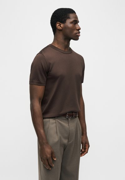 Άνδρας που φοράει εφαρμοστό καφέ πουκάμισο με κοντά μανίκια, κλεισμένο μέσα σε ψηλόμεση μπεζ παντελόνα με πιέτες και πλεκτή καφέ ζώνη.