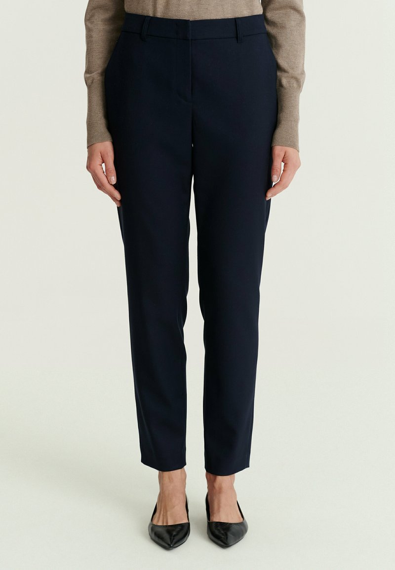 Femme portant un pantalon noir ajusté jusqu’à la cheville, un haut beige à manches longues et des ballerines noires à bout pointu, debout devant un fond uni.