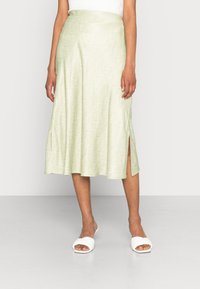 Žena v světle zelené midi sukni s bílými abstraktními liniemi a bočním rozparkem, doplněná bílými sandály na klínu a bílým topem.