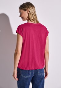 Krátký rukáv, fuchsiové tričko s kulatým výstřihem a nařaseným lemem, spárované s modrými denimovými džíny. Tkanina se zdá být měkká a jemná.