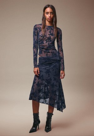 Marineblaues durchsichtiges Kleid mit floraler Stickerei, langen Ärmeln, gerafftem Oberteil und asymmetrischem Saum, getragen mit schwarzen Stiefeletten mit Absatz.