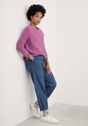 Lila gerippter Pullover mit weiß-blauen gestreiften Bündchen, kombiniert mit lockeren blauen Jeans, weißen Sneakers und mehrfarbig gestreiften Socken.