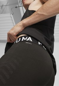 Top deportivo negro sin mangas con tejido texturizado, que presenta una cinturilla con logotipo visible y etiqueta dryCELL. Primer plano de la cintura y la parte superior del muslo.