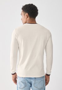 Camisa de manga larga en algodón color off-white con cuello redondo y acentos en burdeos en los puños, combinada con jeans de mezclilla azul claro.