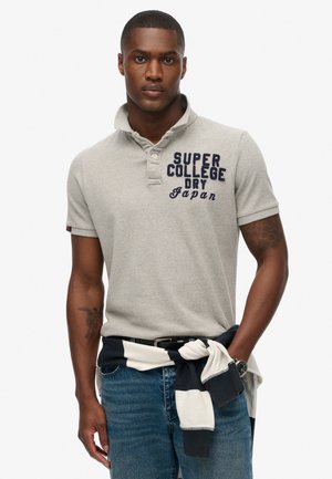 Grå polo skjorte med ribbet krave og korte ærmer, med marineblå trykt tekst. Båret med blå jeans og et marineblå stribet plagg bundet om livet.