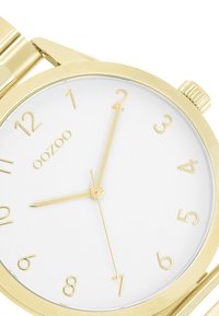 Montre-bracelet en or avec un cadran blanc, présentant des index et des aiguilles en or. La montre est dotée d'un bracelet métallique lisse et d'un boîtier rond.