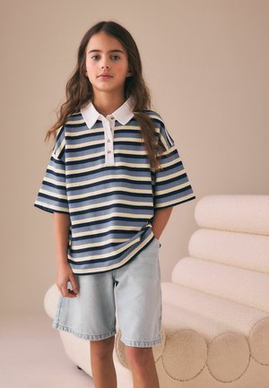 Mergina stovi su ranka kišenėje, vilkinti mėlynos ir baltos spalvų dryžuotą polo marškinėlį ir šviesius džinsinius šortus priešais smėlio spalvos foną.