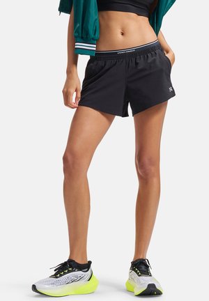 Kobieta w czarnych krótkich sportowych spodenkach z logo Under Armour, białych i neonowo-żółtych butach do biegania, stojąca z rękami w kieszeniach.