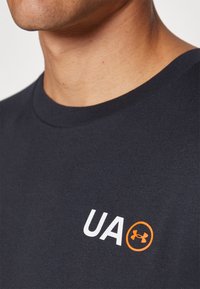 Tume sinine lühikeste varrukatega T-särk, mis on valmistatud puuvillast; vasakul rinnal on valge "UA" tekst ja oranž logo. Klassikaline ümmargune kaelus.