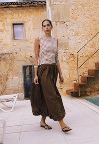 Femme marchant près d'un bâtiment en pierre et d'une piscine, portant un pantalon large marron, un haut sans manches à carreaux et des sandales noires.
