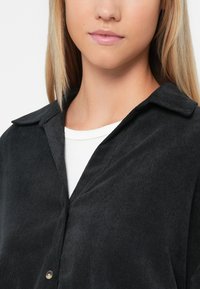 Czarna koszula z kordurojowej tkaniny z dopasowanym kołnierzykiem, z zapięciem na guziki z przodu oraz białą koszulką z okrągłym dekoltem pod spodem, o gładkiej fakturze.