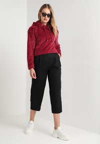 Burgundska fluffy dukserica s kapuljačom i vezicom, u kombinaciji s labavim crnim skraćenim hlačama i bijelim debelim tenisicama. Cool, teksturirani materijal kroz cijelu odjeću.