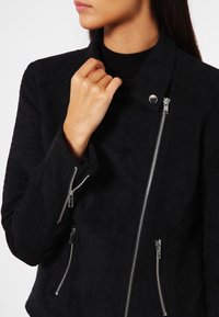 Veste en daim noir avec une silhouette ajustée. Présente des zips en argent et une fermeture à pression. Surface texturée avec des poches zippées sur les côtés.
