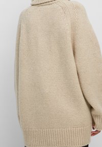 Beige neulepaita, jossa on väljähkö malli ja korkea kaulus. Hihansuut ja helma ovat resorilliset ja teksturoidut. Ei näkyviä koristeita.