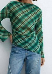 Donna che indossa un top asimmetrico a maniche lunghe a scacchi verdi e jeans blu, in piedi con una mano sui fianchi contro uno sfondo bianco.