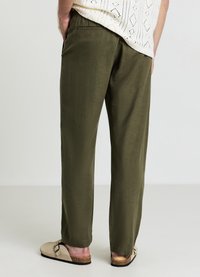 Olivgrüne Hose mit lockerem Schnitt, aus einem weichen Stoff gefertigt. Verfügt über eine einzelne Gesäßtasche und wird mit hellen Slip-On-Schuhen getragen.