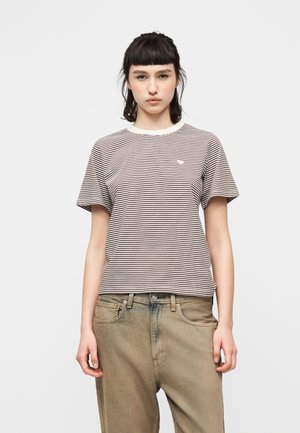 Junge Frau mit kurzen dunklen Haaren, die ein kurzärmeliges, schwarz-weiß gestreiftes Shirt und locker sitzende, ausgewaschene braune Jeans trägt.