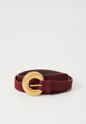 Burgundy suede belt with a large gold circular buckle. Features a smooth texture and a slim design, secured with a matching strap loop.