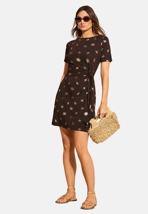 Kobieta w czarnej sukience z krótkim rękawem z nadrukiem złotych muszelek, okulary przeciwsłoneczne, złote sandały na paski oraz trzymająca teksturowaną beżową torebkę.