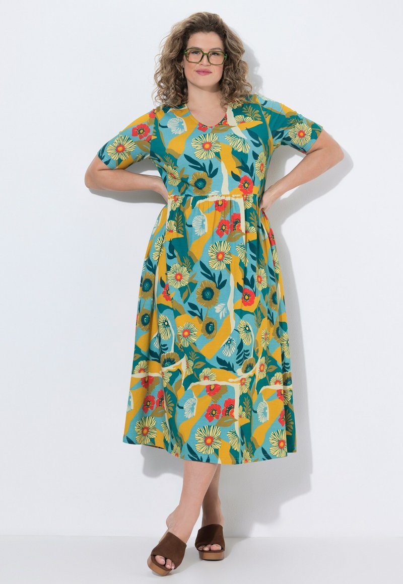 Žena s kudrnatými vlasy a brýlemi, oblečená v barevných květovaných šatech střední délky a hnědých sandálech s otevřenou špičkou, stojící s rukama v bok.