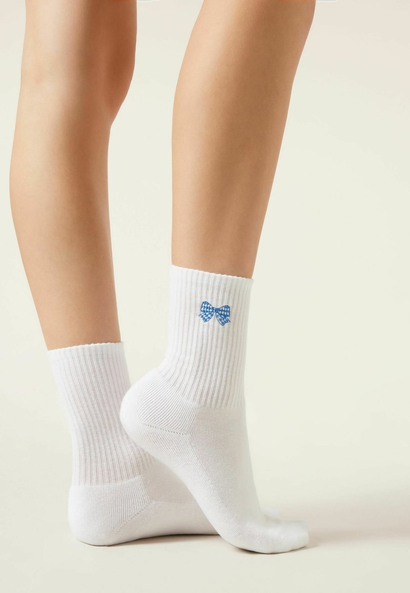 Beine, die weiße gerippte Knöchelsocken mit einer kleinen blauen Schleife auf der Außenseite tragen, stehen vor einem neutralen Hintergrund.