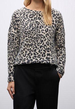 Mujer con un suéter difuso de estampado de leopardo y pantalones negros, de pie con una mano en el bolsillo contra un fondo liso.