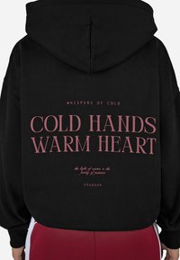 Čierna mikina s kapucňou so veľkým ružovým grafickým textom na zadnej strane, ktorý znie "STUDENÉ RUKY, TEPLÉ SRDCE." Text obsahuje ďalšie citáty a názov značky.