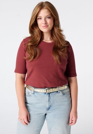 Vrouw met lang golvend rood haar, een roestkleurig shirt met korte mouwen, lichtblauwe spijkerbroek en een metalen gevlochten riem, staand tegen een witte achtergrond.
