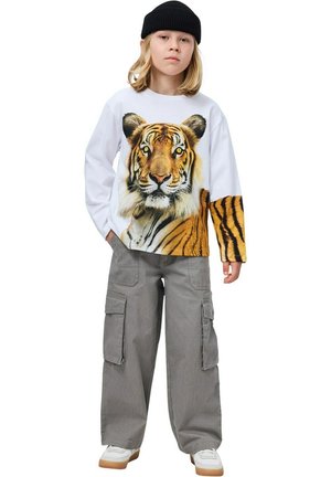 Wit shirt met lange mouwen met een tijgerafbeelding en oranje strepen, gecombineerd met grijze cargobroek en witte sneakers. Zwarte muts op het hoofd.