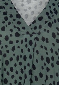 Zelená tkanina s výstřihem do V, která má černý vzor leopardího tisku. Hladký povrch s různými velikostmi a tvary teček.