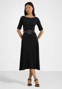 Black, long-sleeve dress with a boat neckline, cinched waist by a large buckle belt, flowing silhouette, worn with black heeled sandals.
