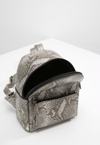 Mochila gris con un patrón y textura de piel de serpiente, que cuenta con una apertura con cremallera, correas ajustables y un bolsillo delantero.